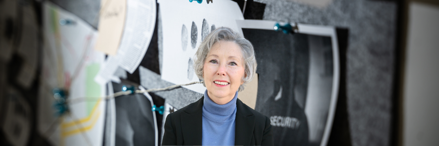 An older woman in a blue sweater and black jacket is smiling in front of a wall.