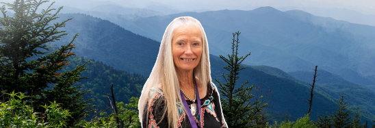 A woman with long blonde hair is standing in front of a mountain range.