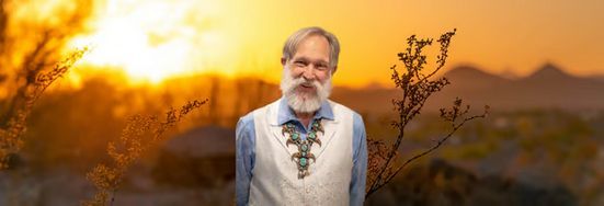 An older man with a beard is standing in front of a sunset.
