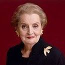 An older woman wearing a black jacket and a brooch is standing in front of a red background.