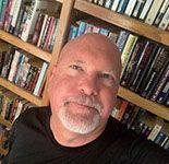 A bald man with a beard is taking a selfie in front of a bookshelf filled with books.