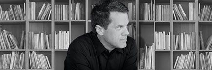 A man in a black shirt is sitting in front of a bookshelf.