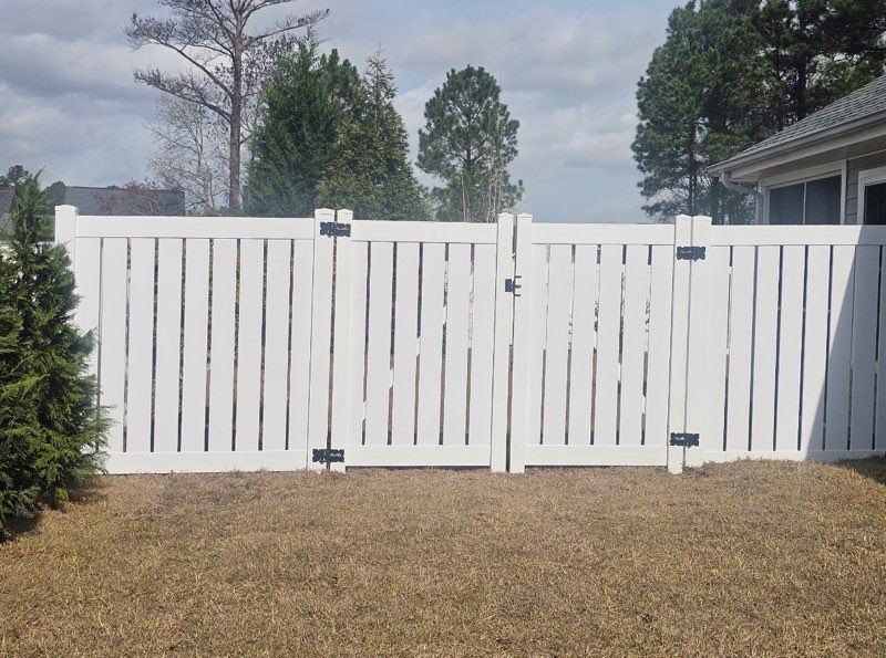A white vinyl privacy fence with a double gate in a backyard setting under a blue sky.