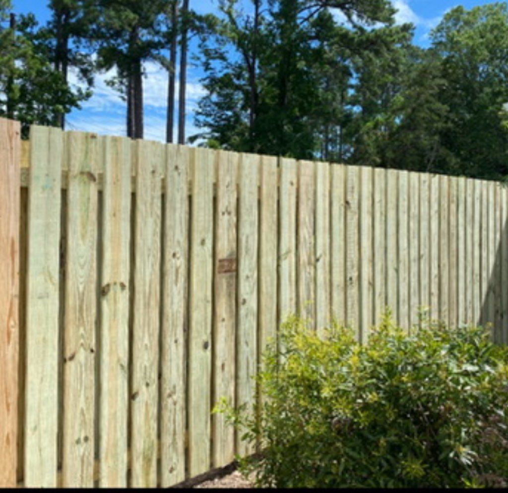 A tall, light-colored wooden privacy fence stands outdoors with green trees and a blue sky in the background.