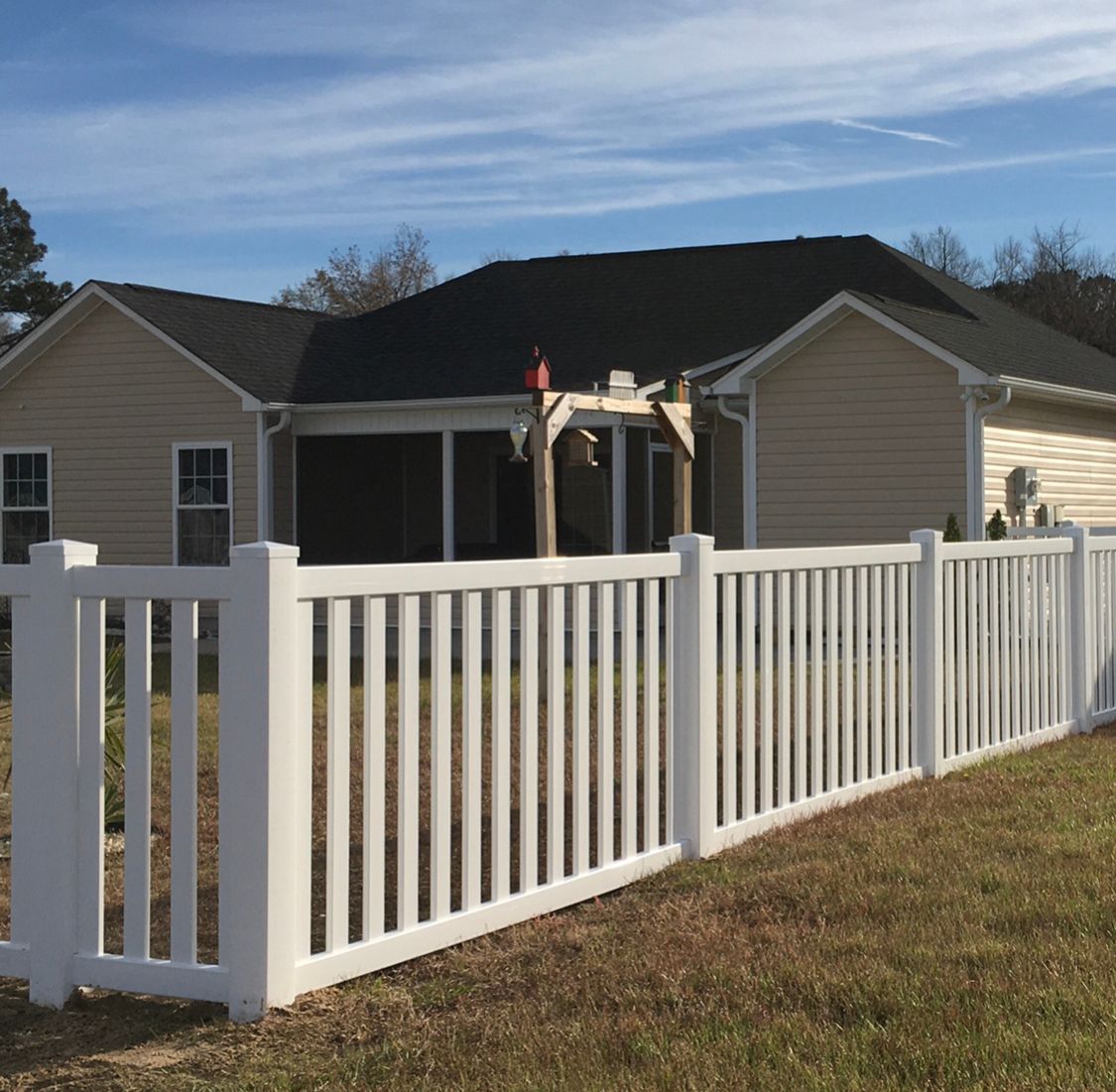 A beige house with a black roof and a screened-in porch sits behind a white vinyl fence in a grassy yard under a blue sky.