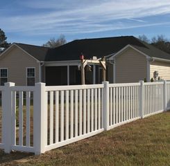 A beige house with a dark roof and a screened porch is enclosed by a white vinyl fence in a grassy yard under a blue sky.