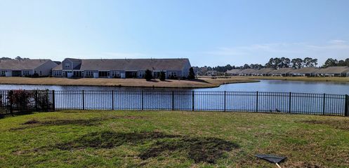 A view across a grassy yard and a dark fence toward a calm lake lined with suburban houses under a clear blue sky.