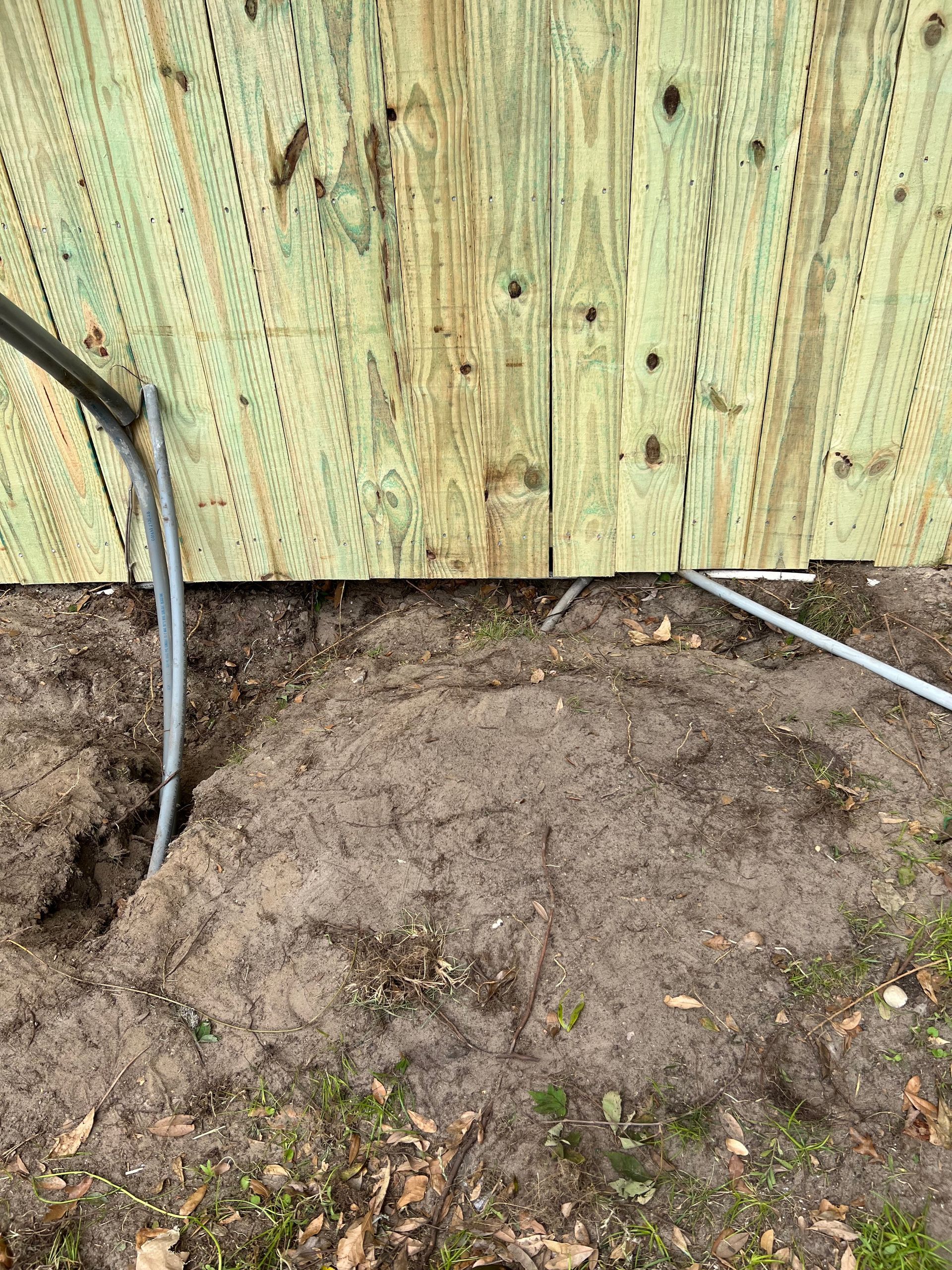 Two gray electrical conduits emerge from the ground and run up the base of a light-colored wooden fence.