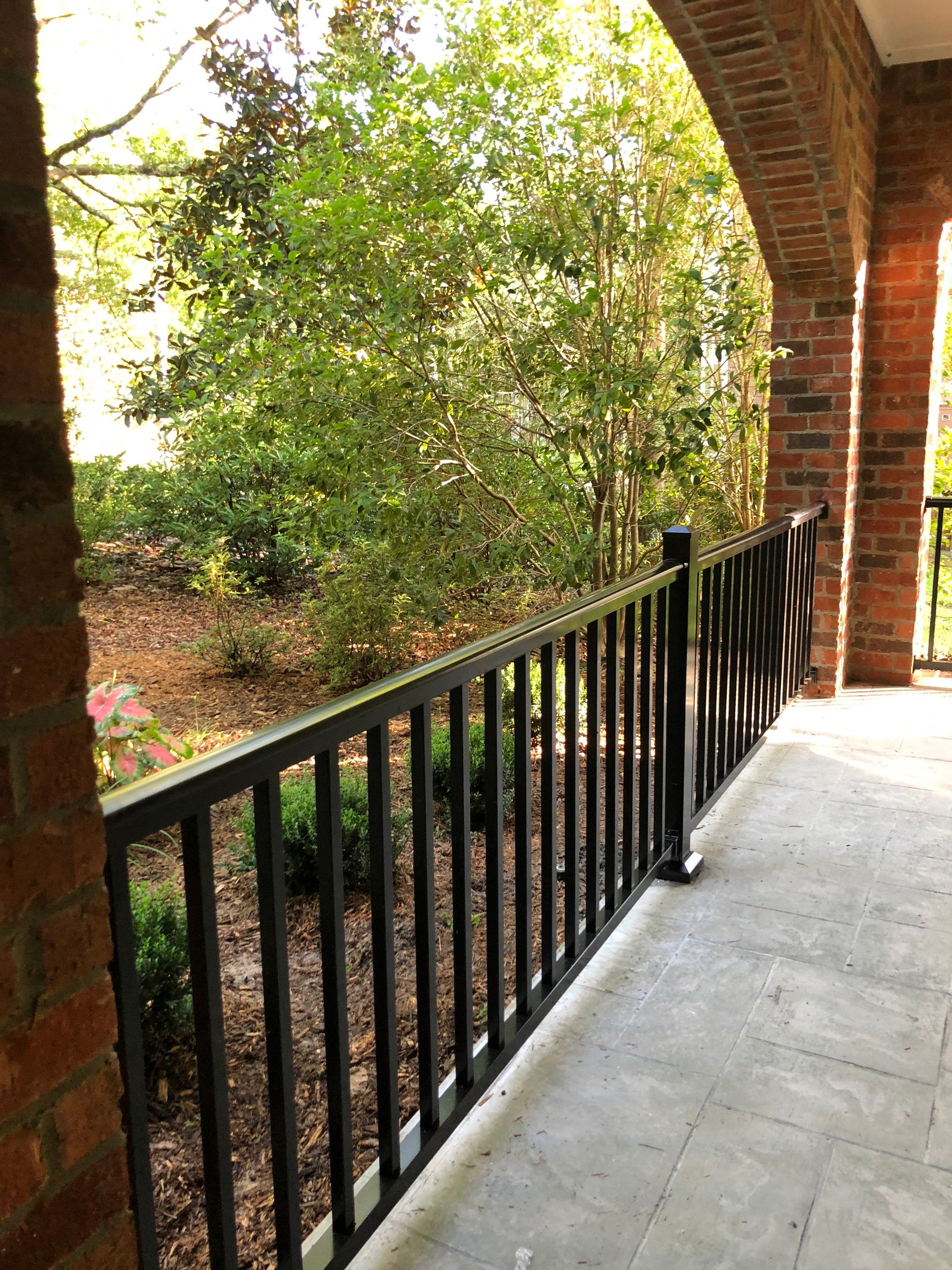 Black metal railing on a stone porch overlooking a garden with trees and shrubs.