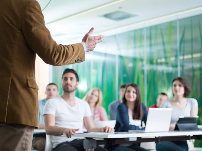 Il relatore si rivolge a un pubblico seduto in una sala conferenze con computer portatili e uno sfondo verde.