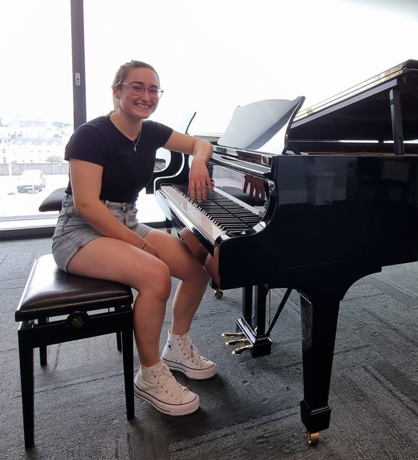 Person seated at a grand piano, smiling in a bright room with large windows.