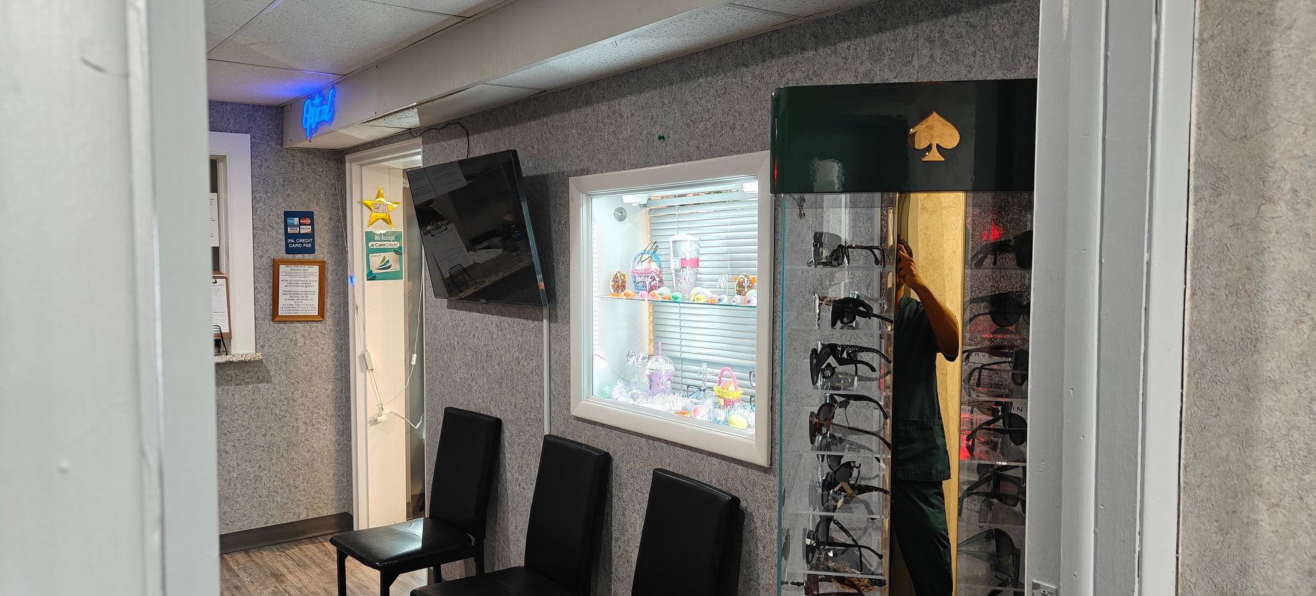 An eyeglass store waiting area with display cases, seating, and a wall-mounted television.