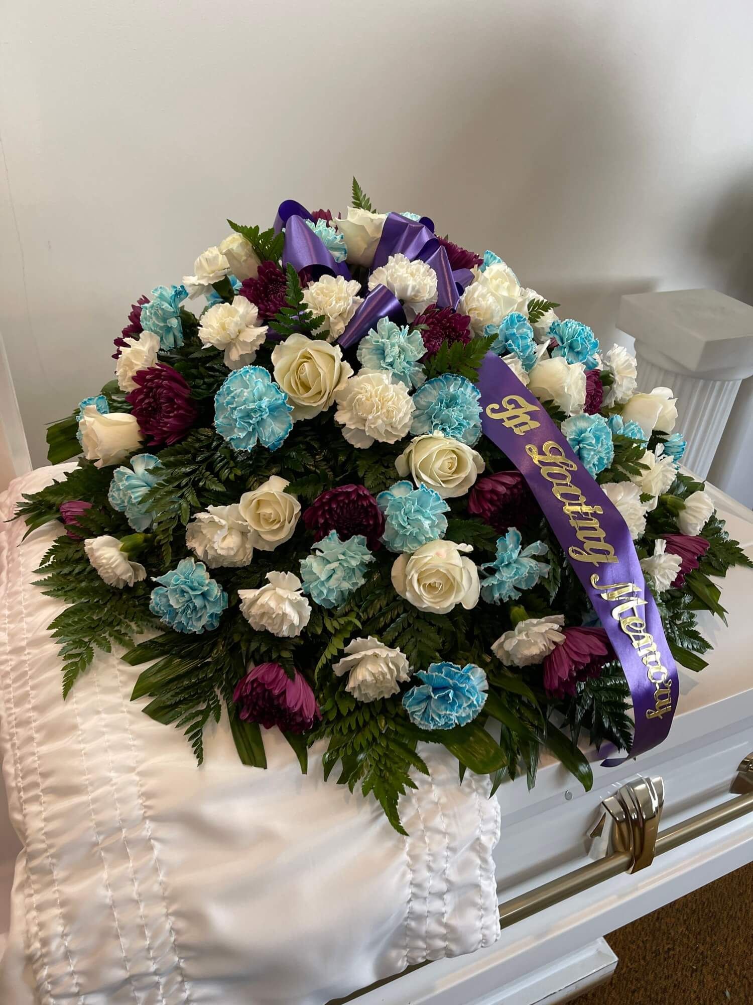 White floral funeral spray with purple ribbon resting on a white casket inside a room