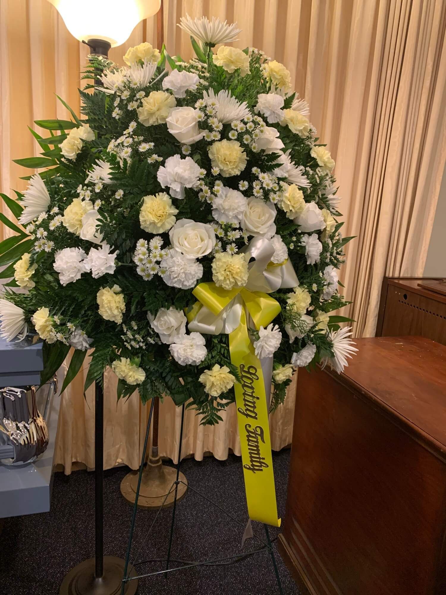 White floral funeral wreath with yellow ribbon on an easel indoors, against beige curtains.