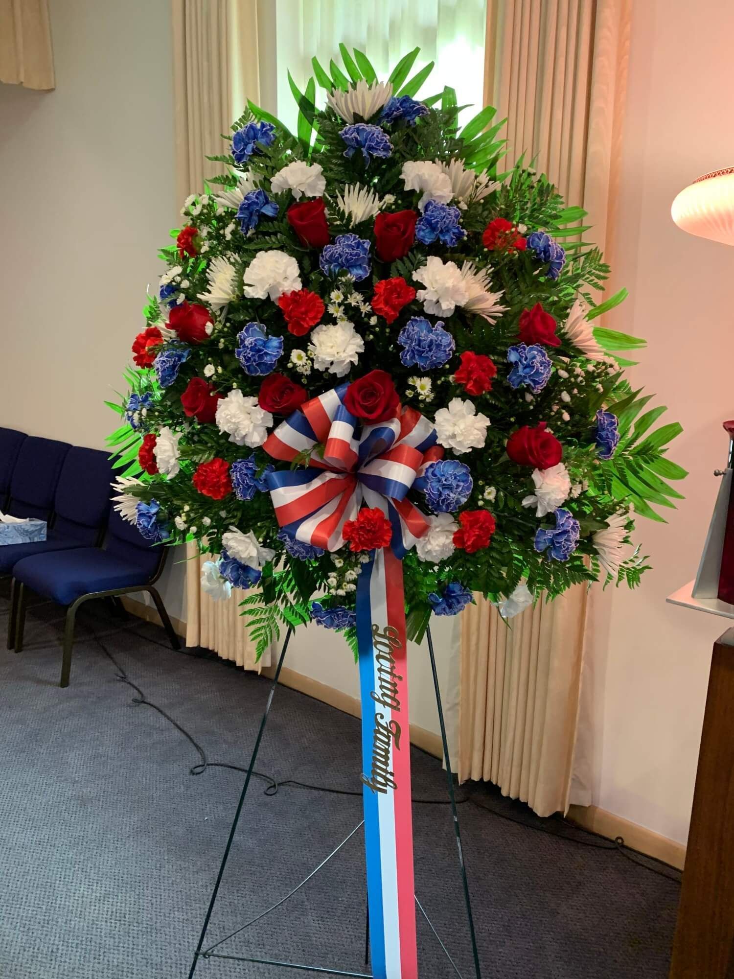 Floral wreath in red, white, and blue on a stand with ribbon in a room.
