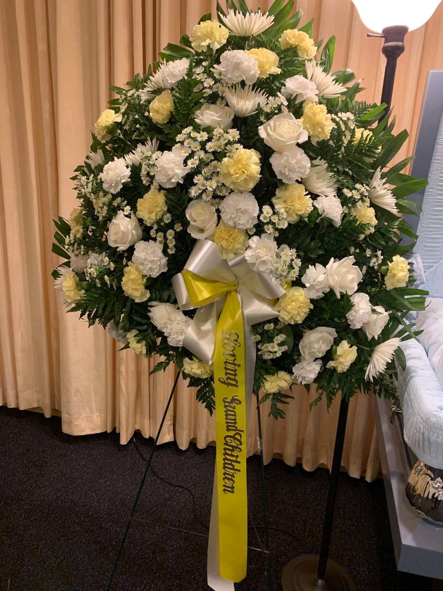 White and yellow funeral wreath with yellow ribbon on an easel in front of beige curtains