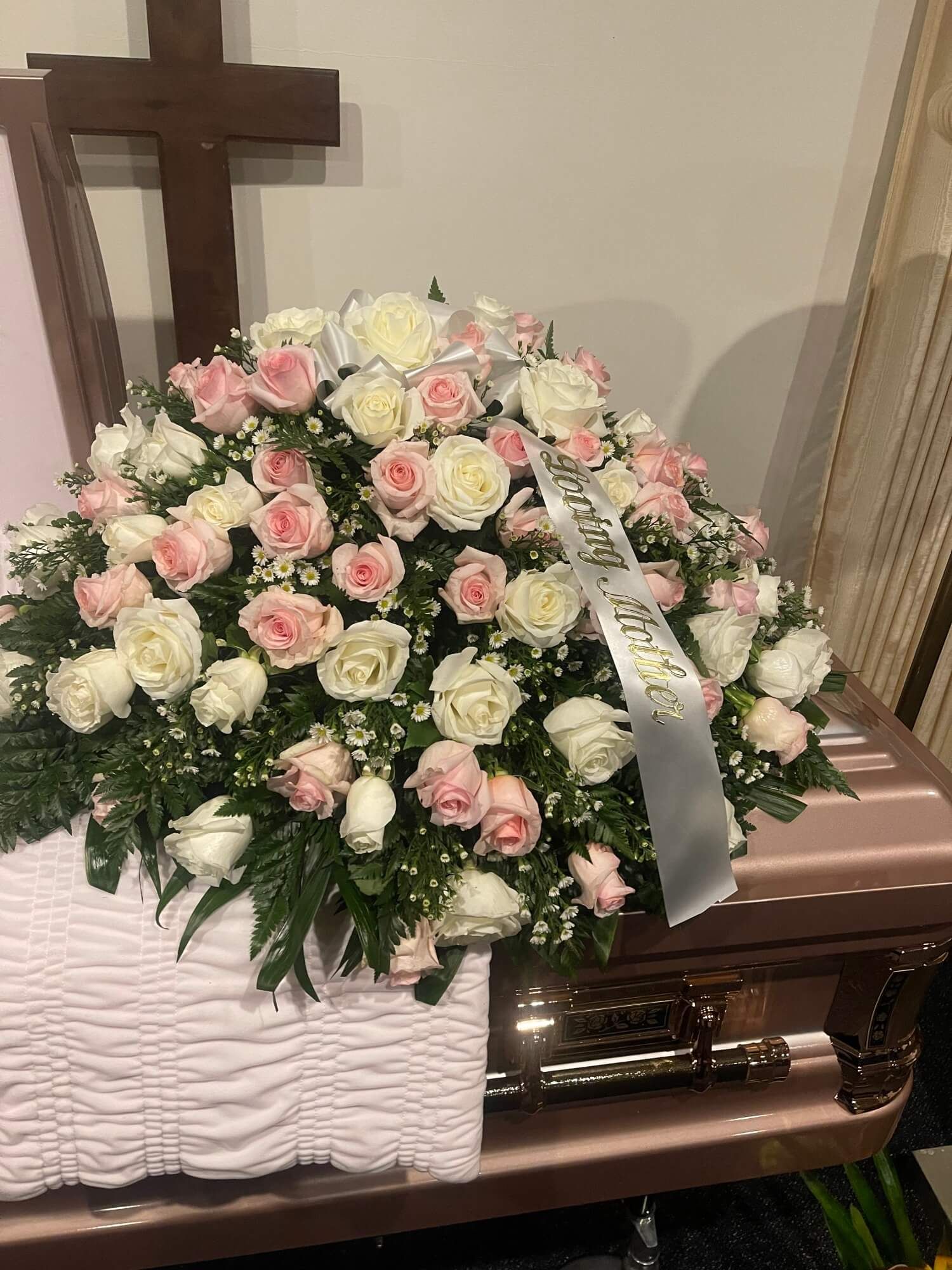 Pink and white rose casket spray on a brown casket with a white ribbon and cross backdrop