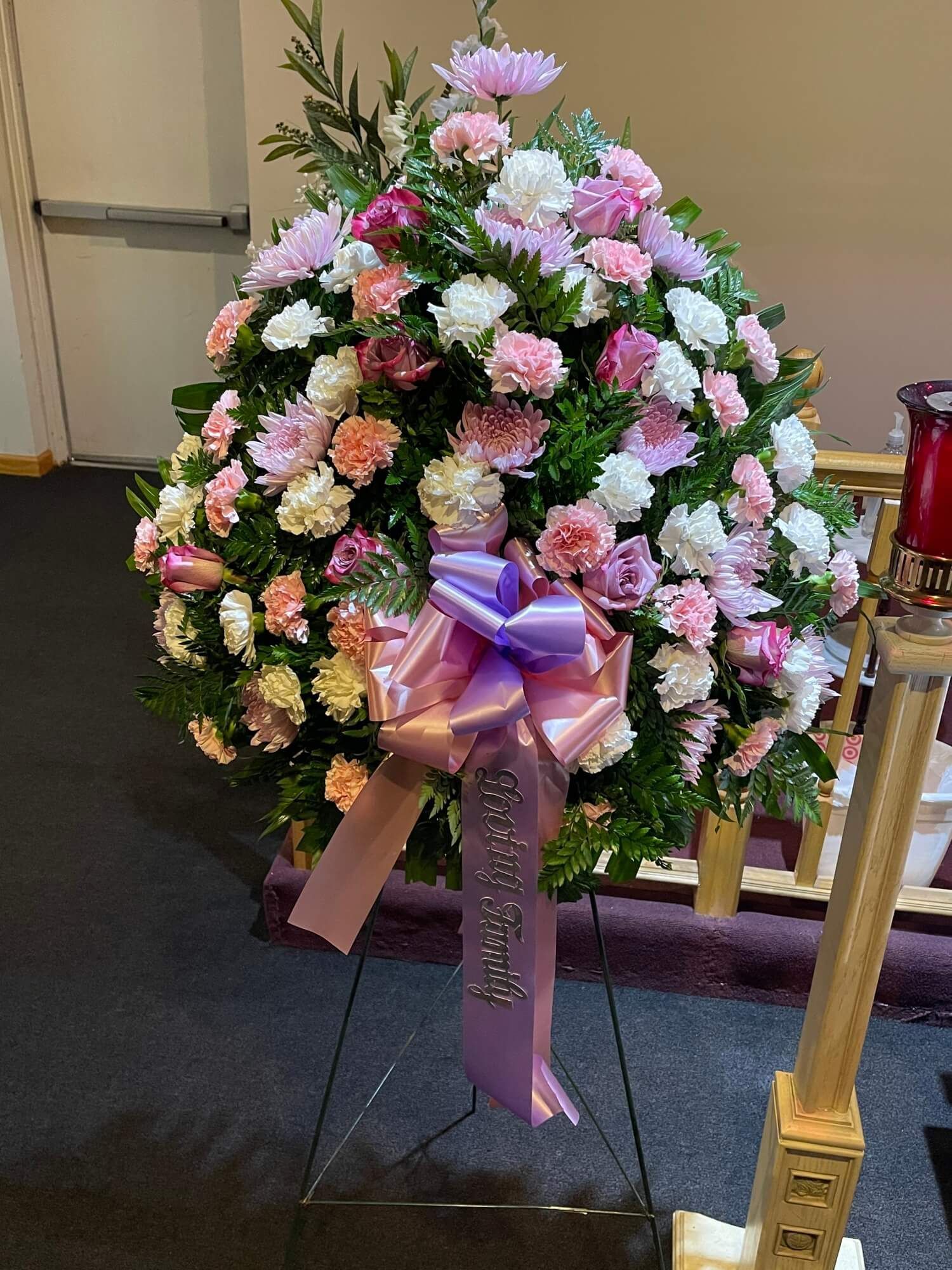 Pink and white floral funeral wreath with purple ribbon on a stand indoors
