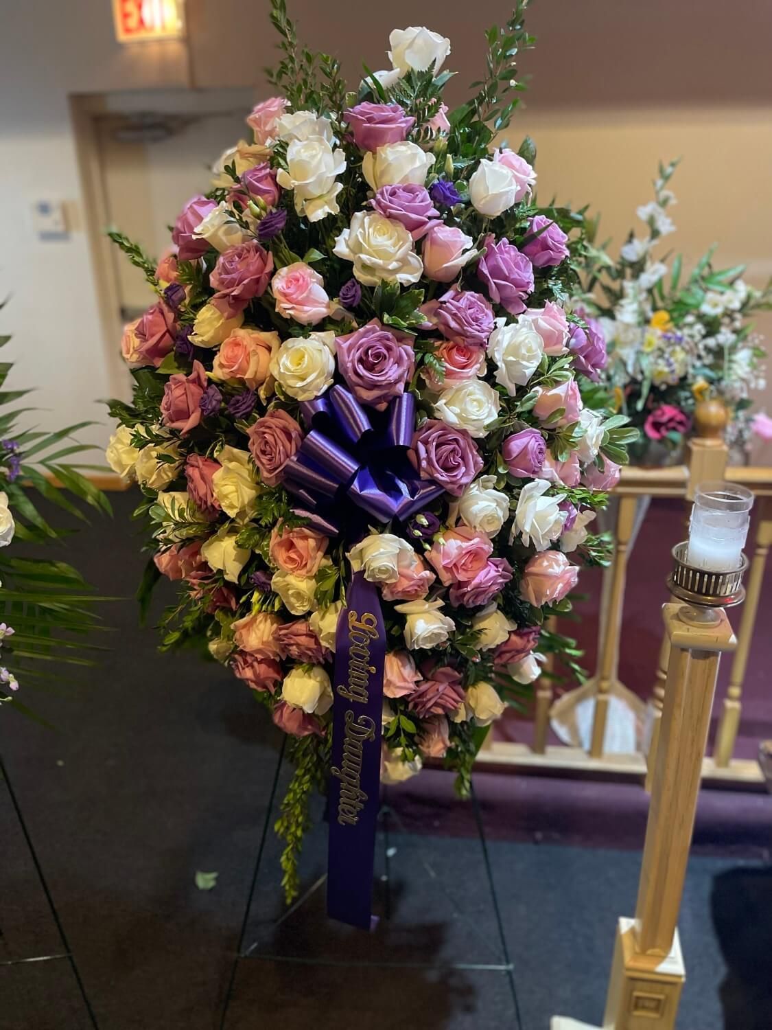 Large floral spray of pink, purple, and white roses with a purple ribbon on an easel indoors