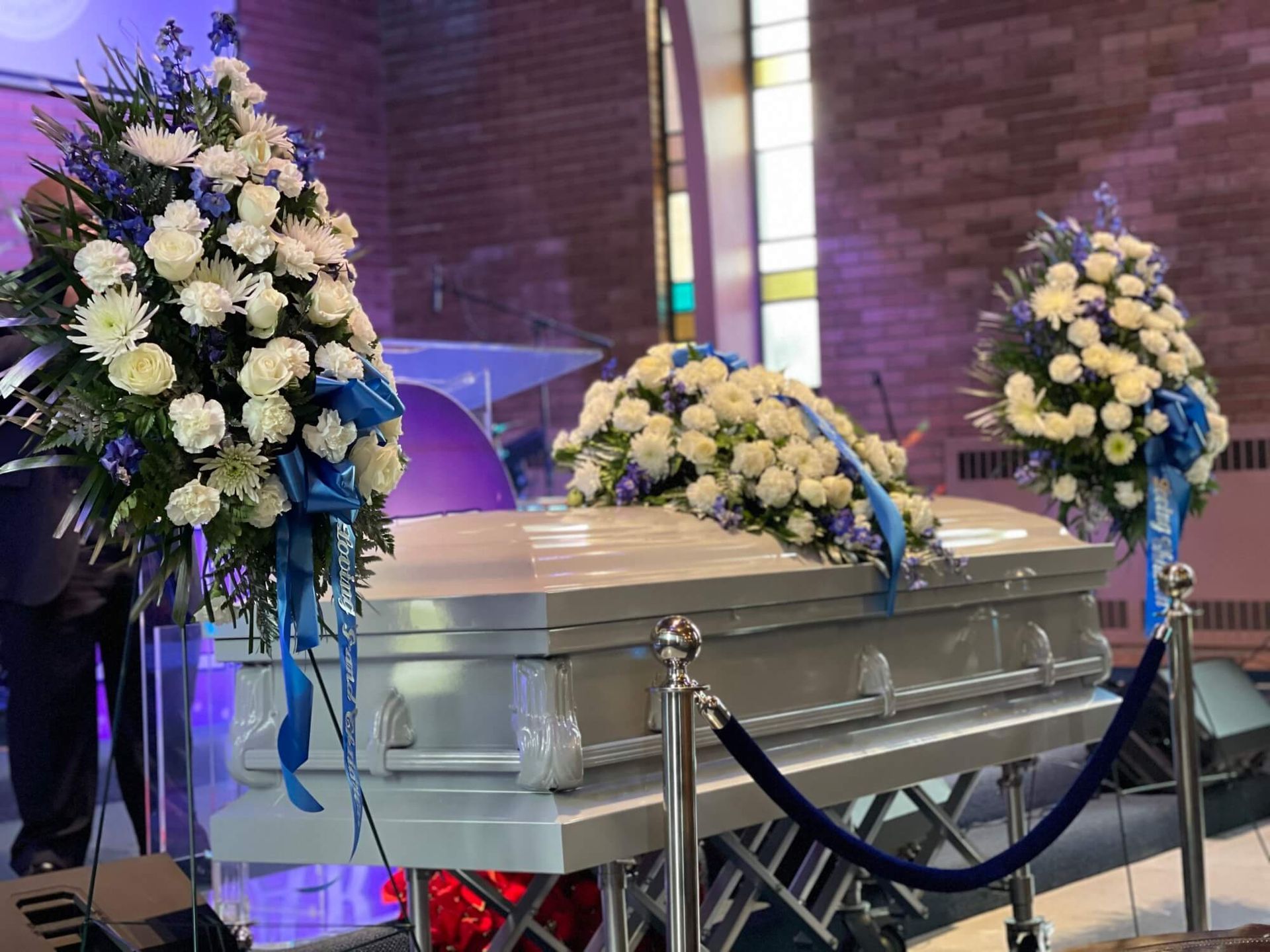 White casket with blue and white floral arrangements in a church chapel, framed by velvet ropes
