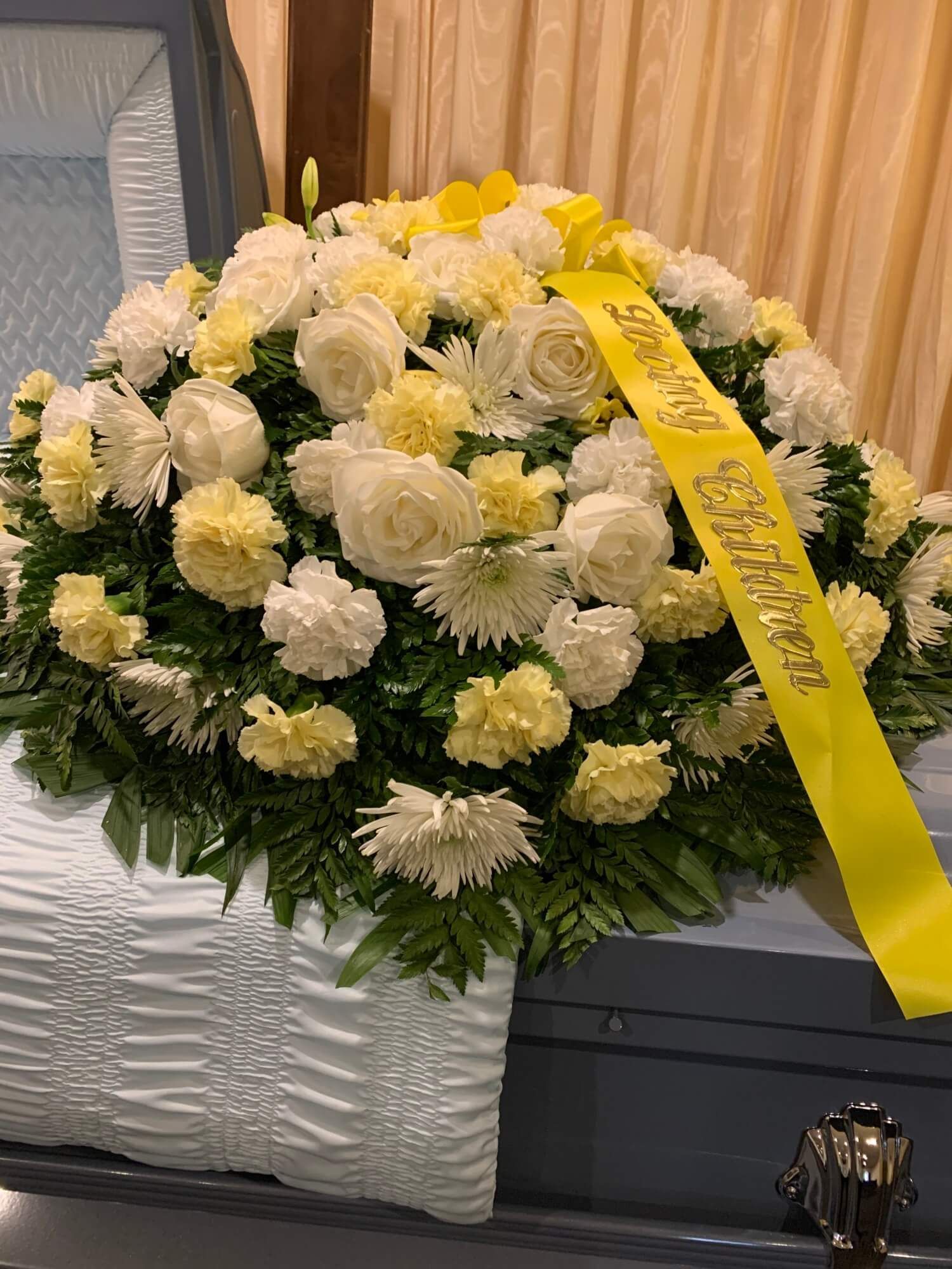 White and yellow floral funeral wreath with a yellow ribbon, resting on a casket