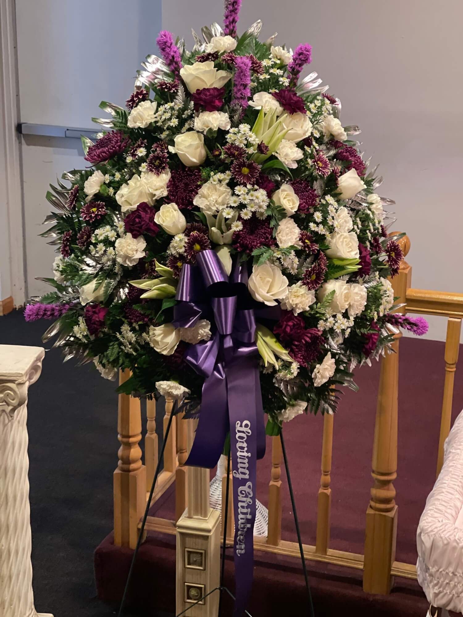 Purple and white funeral wreath on a stand with ribbon in a church or chapel setting