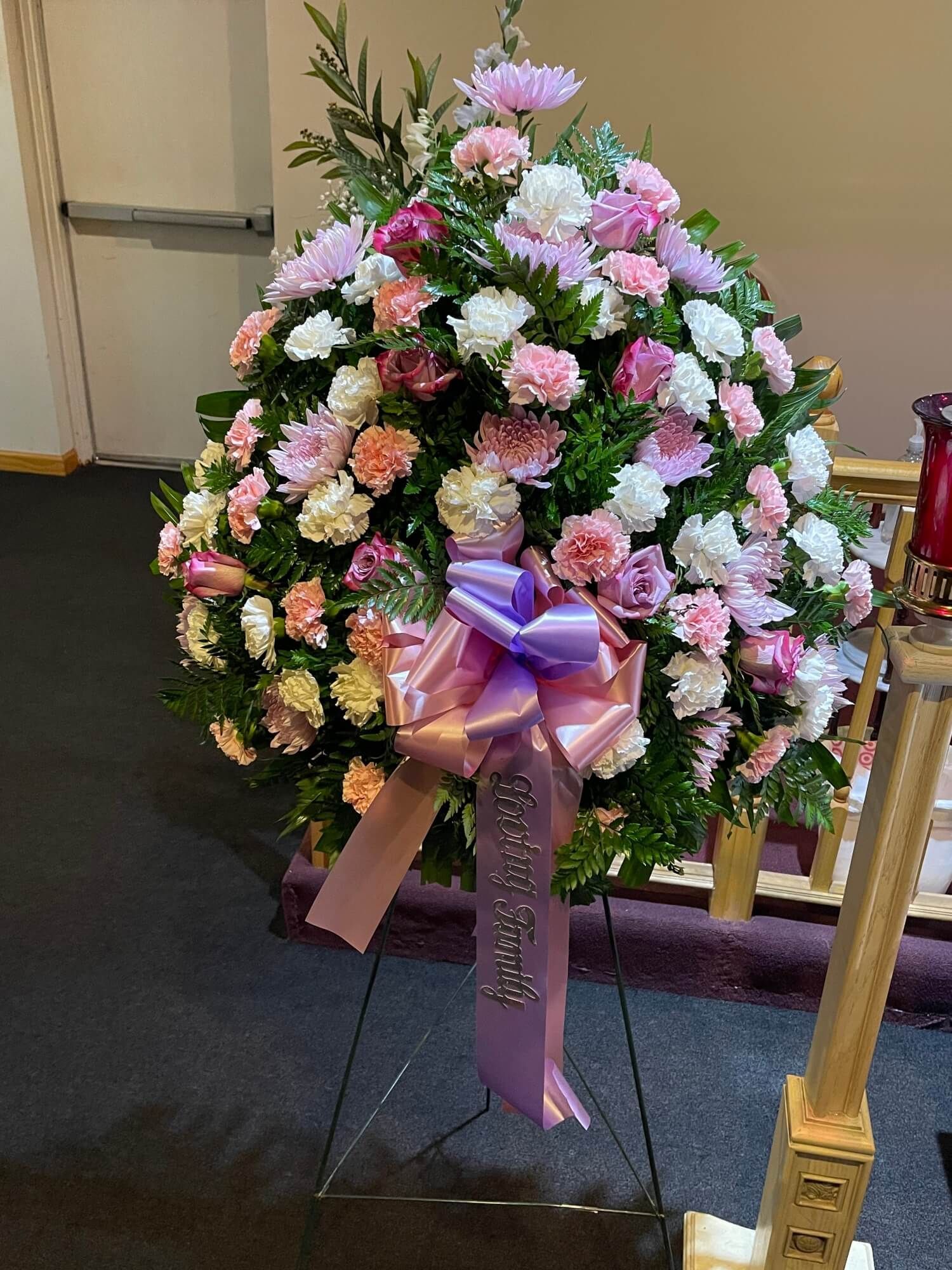 Pink and white floral wreath on a stand with a purple bow in an indoor setting