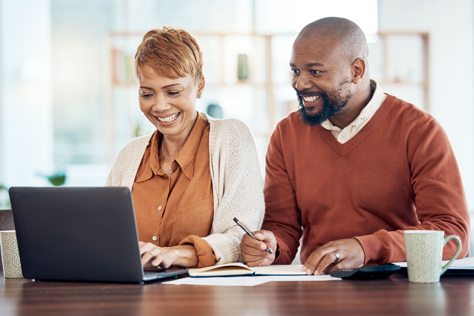 An elderly couple is sitting at a table with a laptop and a notebook.