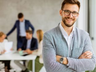Ein lächelnder Mann mit Brille und verschränkten Armen sitzt in einem Büro; im Hintergrund sind seine Kollegen verschwommen zu sehen.