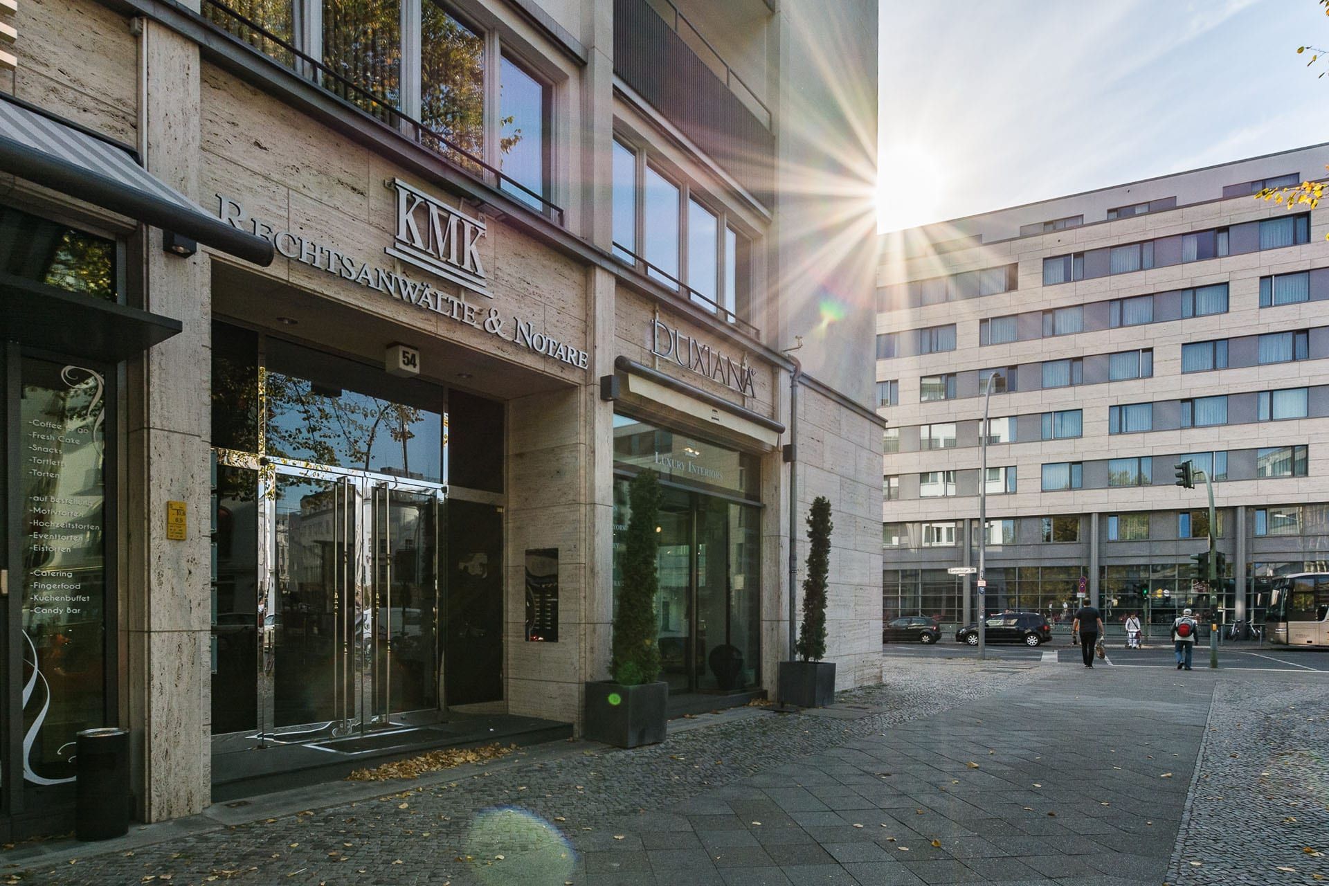Sonnendurchfluteter, moderner Gebäudeeingang mit Glastüren, die sich zu einem gepflasterten Stadtplatz öffnen