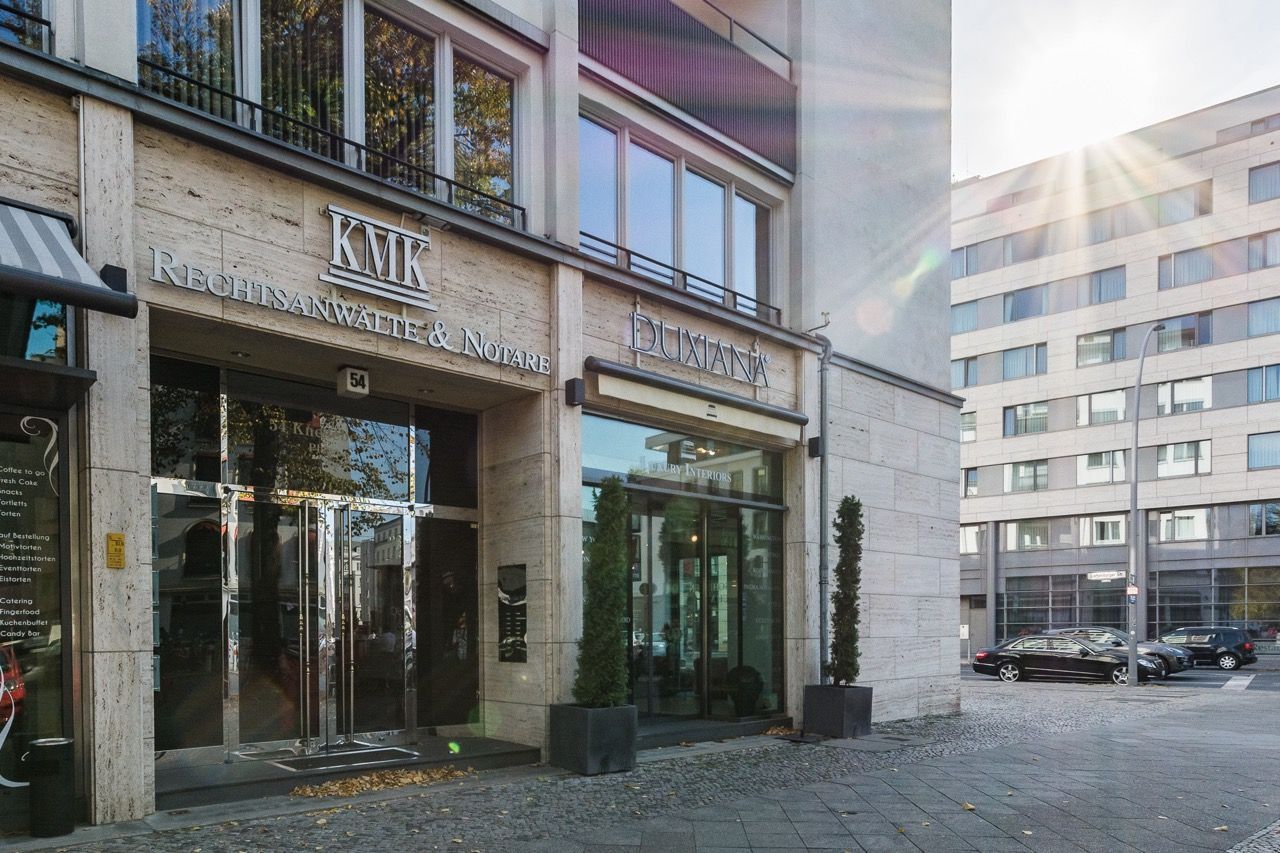 Moderner Gebäudeeingang mit Glastüren an einer Kopfsteinpflasterstraße, Sonnenlicht spiegelt sich in den Fenstern