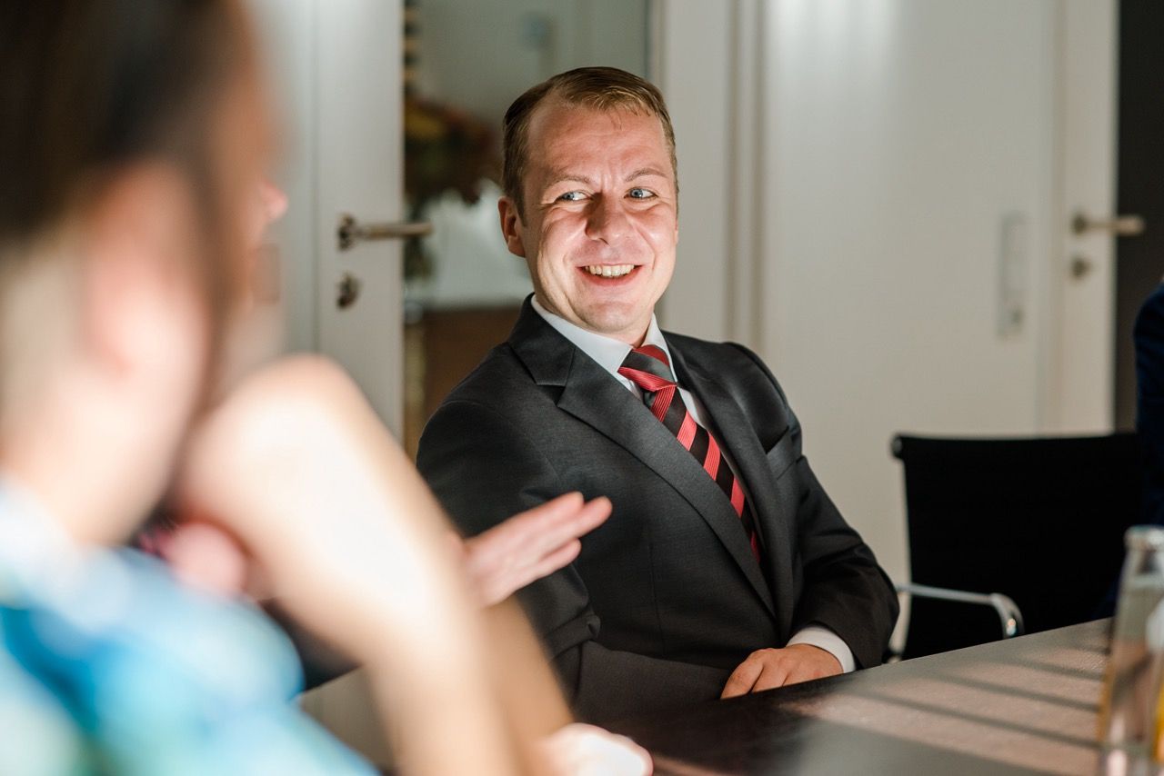 Geschäftstreffen in einem Konferenzraum, ein Mann im Anzug lächelt ihm gegenüber am Tisch zu.