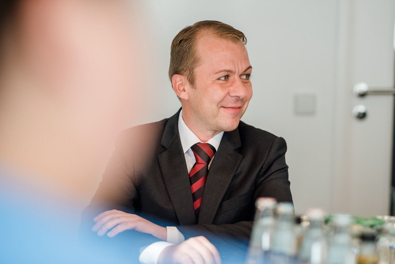 Mann in dunklem Anzug und rot gestreifter Krawatte lächelt an einem Besprechungstisch; im Vordergrund sind verschwommene Glasflaschen zu sehen.