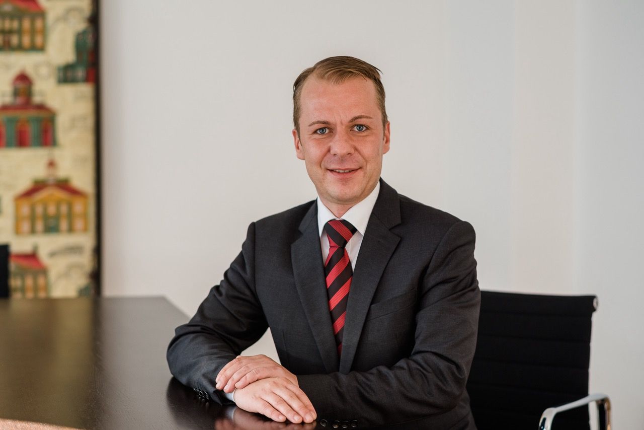 Ein Mann in einem dunklen Anzug und einer rot gestreiften Krawatte sitzt an einem Schreibtisch in einem Büro und blickt in die Kamera.