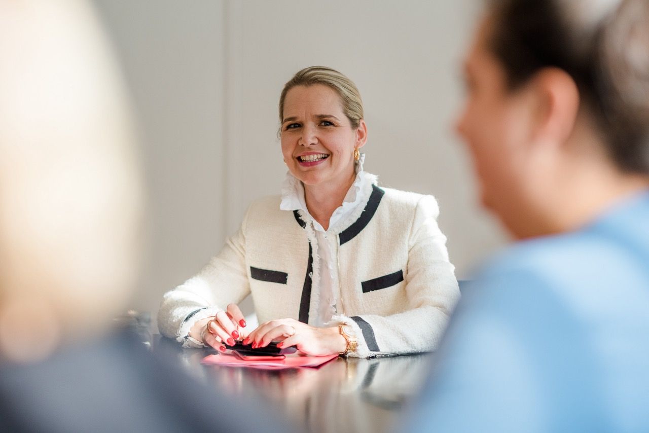 Eine Frau lächelt ihr in einem hellen Besprechungsraum über den Tisch hinweg zu; im Vordergrund sind zwei verschwommene Personen zu sehen.