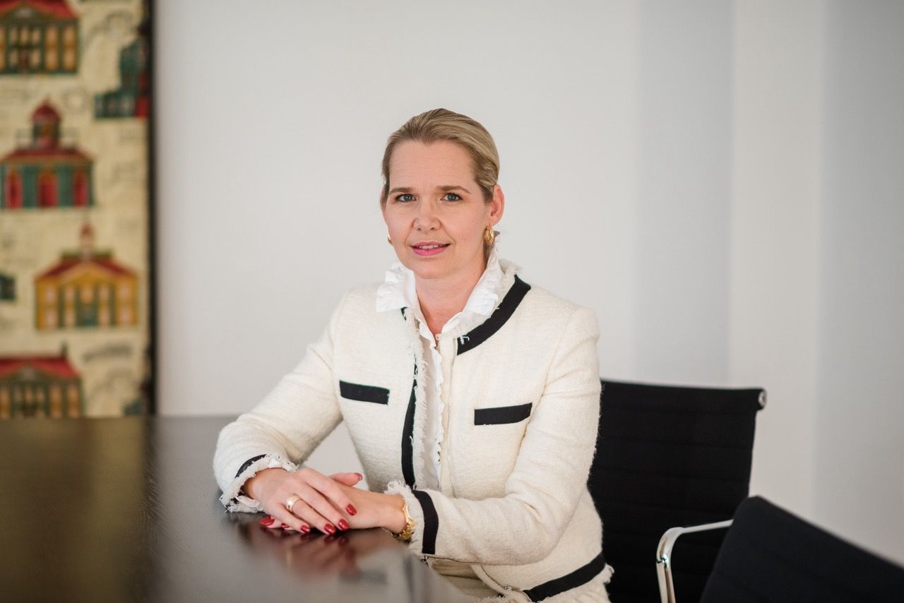 Eine Frau in einem weißen Blazer sitzt lächelnd an einem Konferenztisch in einem hellen Büro.
