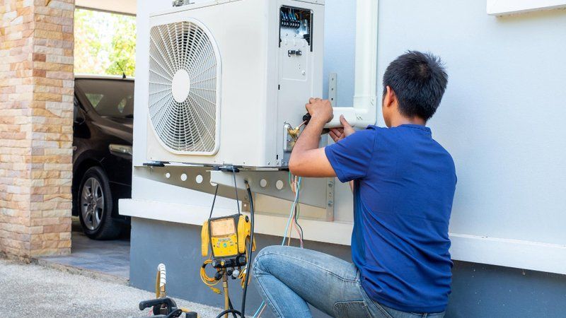 Technician in blue shirt repairing an outdoor air conditioner unit beside a white wall.