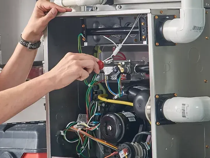 Technician wiring a furnace control panel with open access and visible pipes.