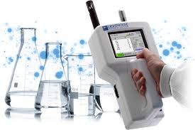 Handheld laboratory meter in gloved hand beside glass beakers and flasks with blue liquid splashes