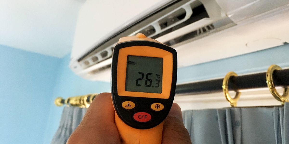 Handheld infrared thermometer aimed at a wall-mounted air conditioner above a curtain rod