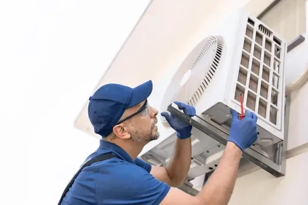 Technician in blue uniform adjusting a wall-mounted air conditioner with a screwdriver