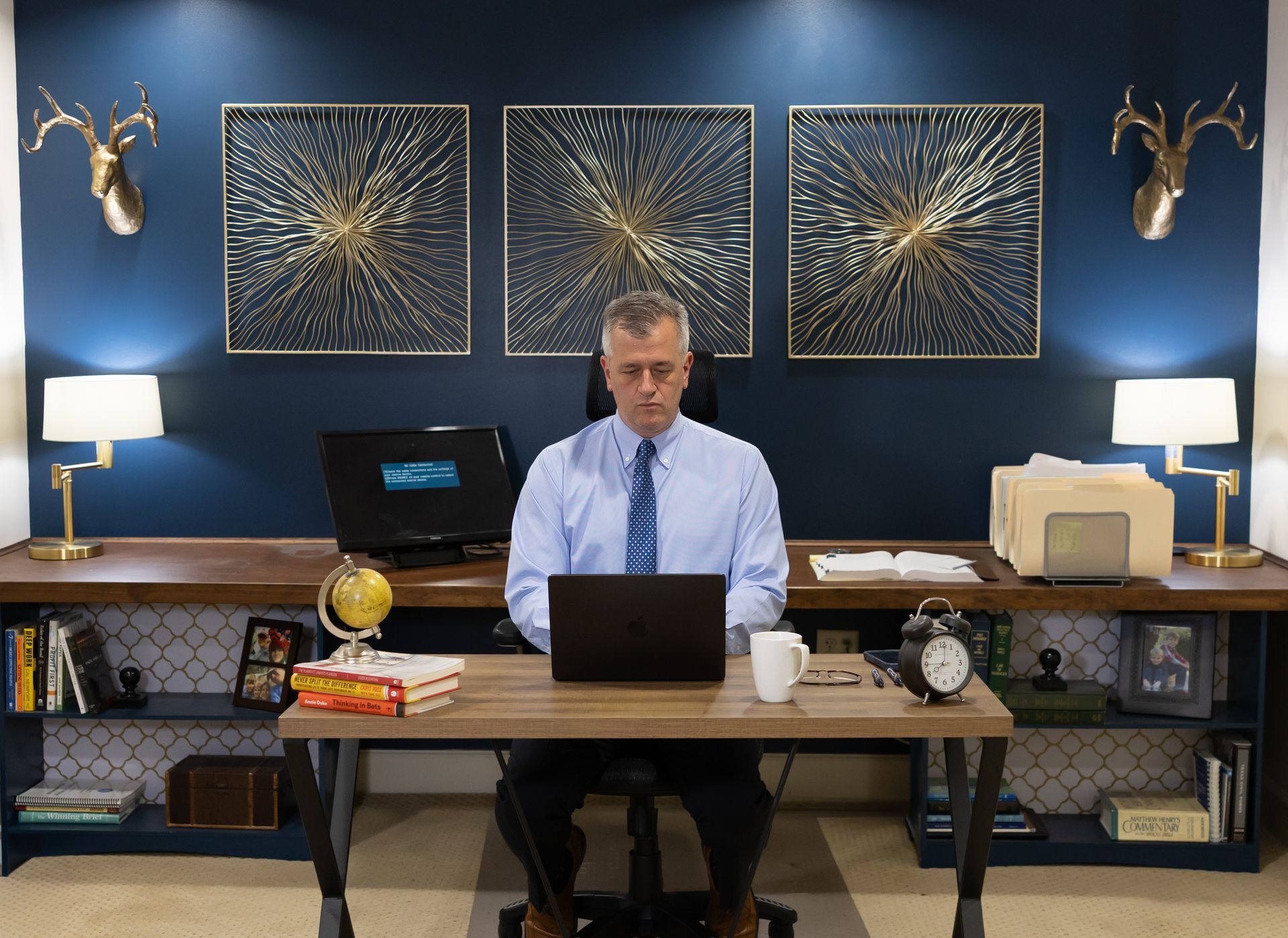Man at desk in blue office, working on laptop between two lamps and framed art.