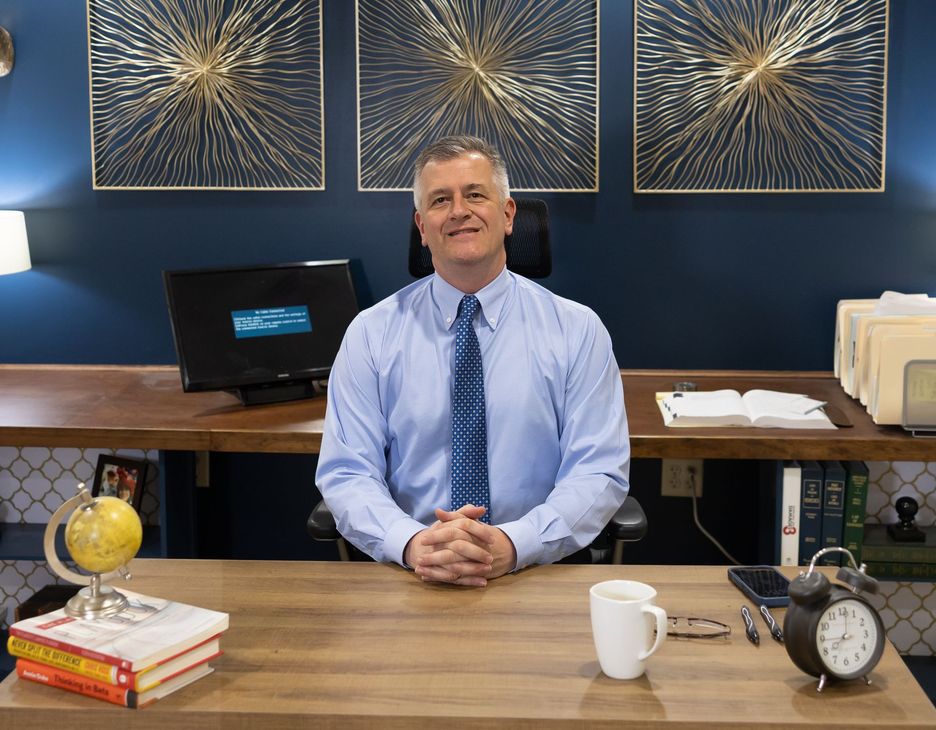 Man seated behind a wooden desk in an office, wearing a light blue shirt and tie, smiling at the camera.