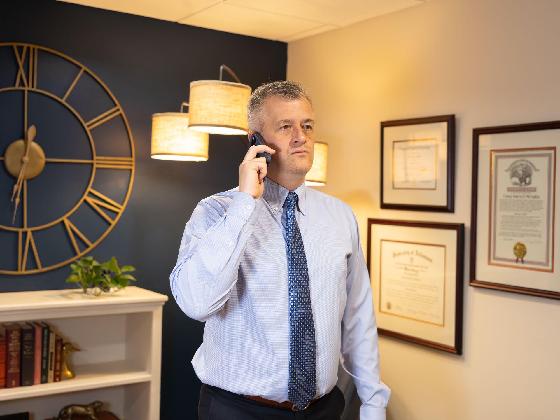 Man in dress shirt and tie talking on phone in an office with framed certificates and a large wall clock
