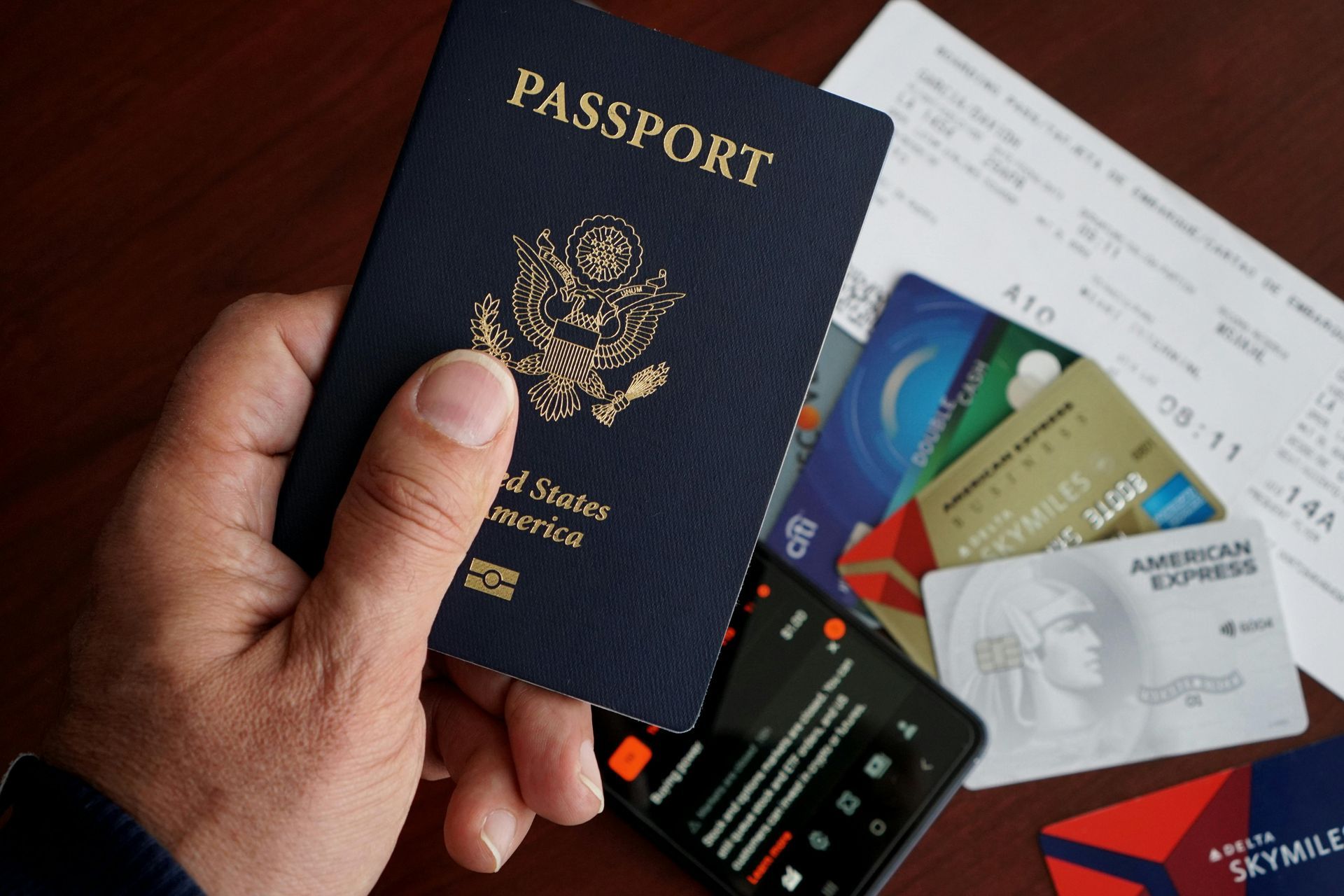 Hand holding a passport beside bank cards, cash, and travel documents on a table