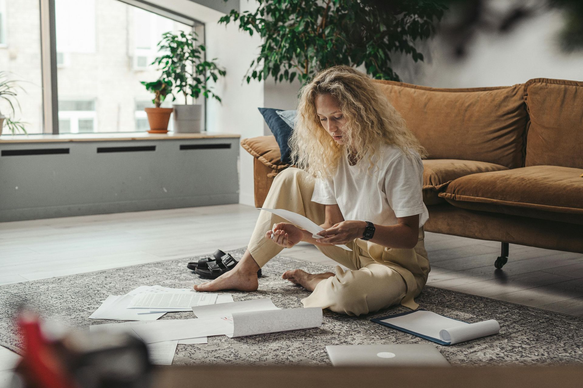 Woman sitting on a rug, working with papers and a laptop in a bright living room.