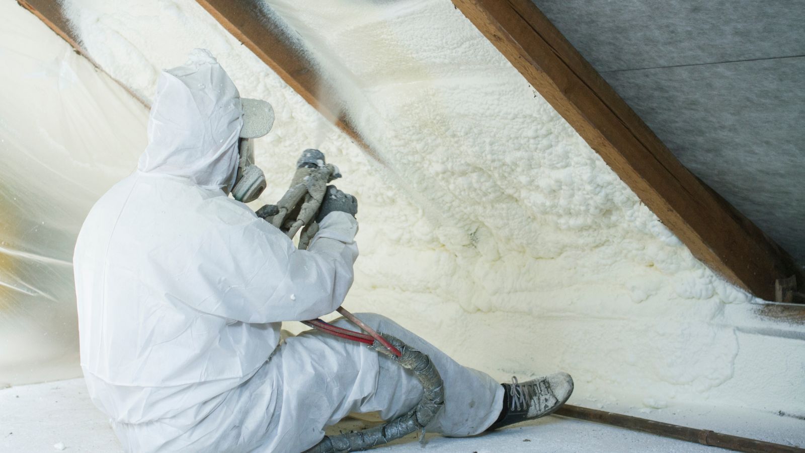 A man in a white suit is spraying insulation in an attic.