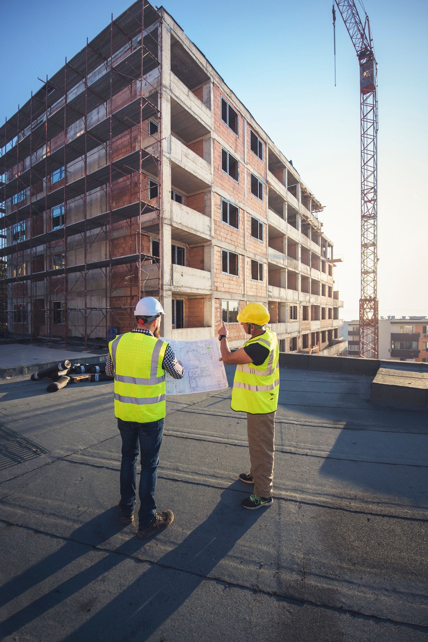 Two construction workers in yellow vests and hard hats inspect a building site beside a partially built brick structure.