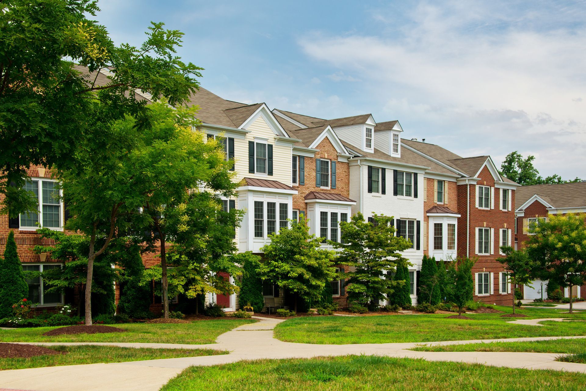 Row of brick townhouses with white trim beside a tree-lined sidewalk and green lawn