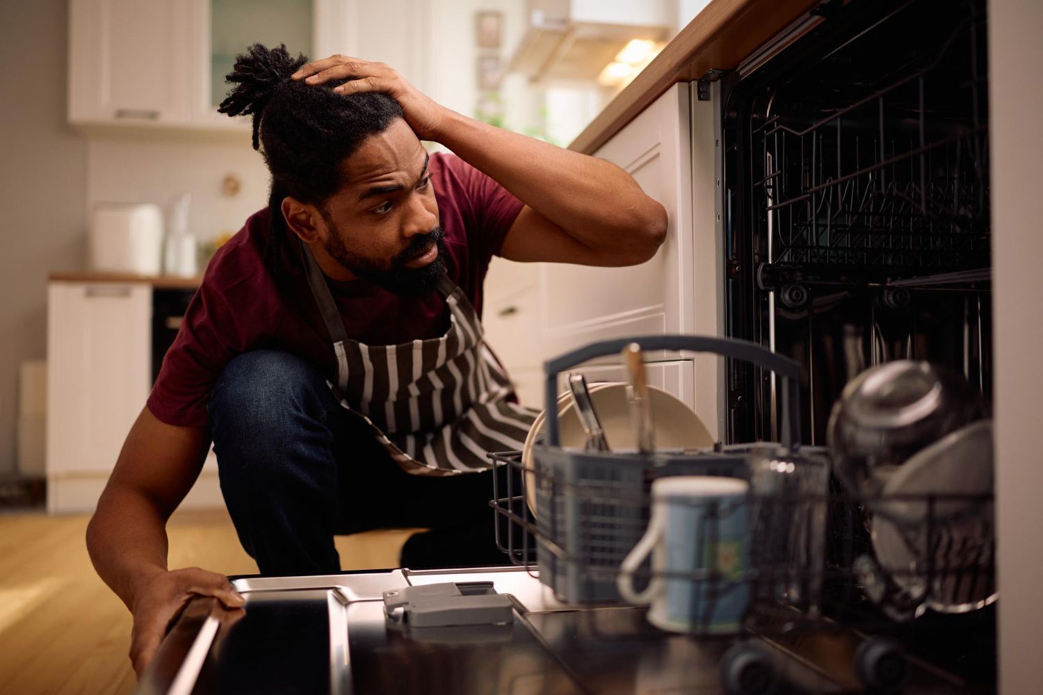 Man in apron, inspecting dishwasher with concerned expression, kitchen background.